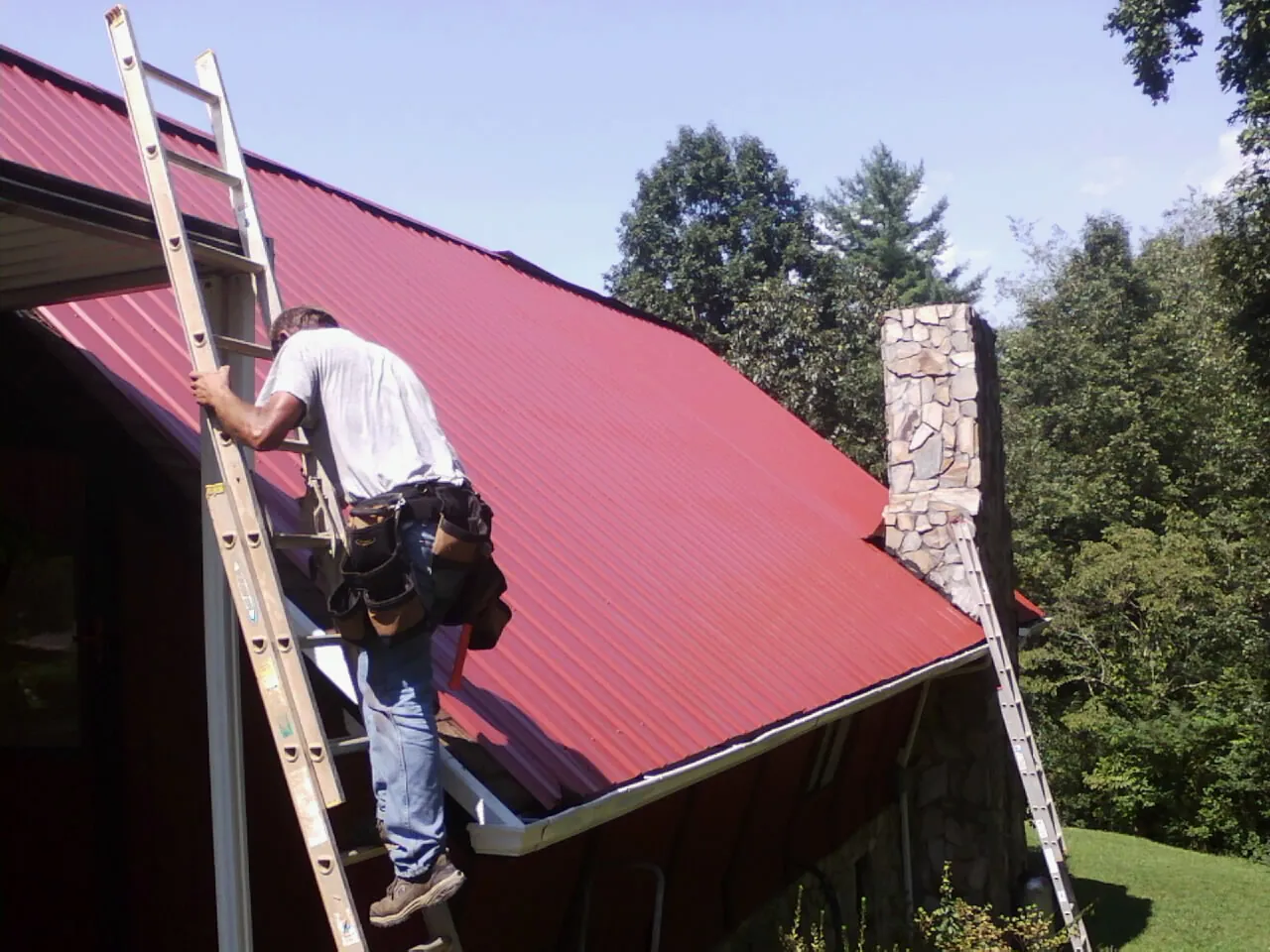 Roofer on a ladder beside a red metal roof and stone chimney — Soffit & Fascia Repair by Roofing Brothers in Cranberry Township